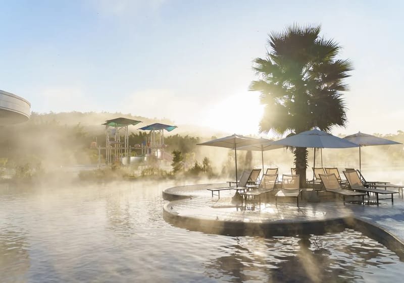 Piscina termal do Jardins de Jurema com vapor ao amanhecer, guarda-sóis e palmeira
